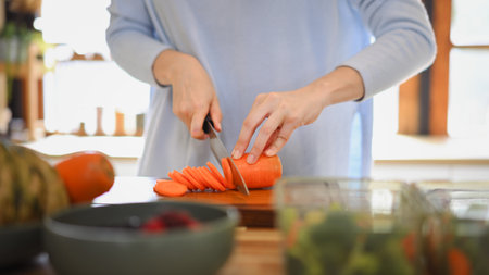 Closeup of young female hands chopping fresh orange carrot on board while in modern kitchen.の写真素材