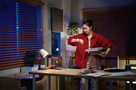 Interior designer checks her watch while holding material samples during late night work in a home office.の写真素材