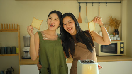 Two young women smiling and holding bread slices together in a cozy home kitchen, enjoying a cheerful cooking moment.の写真素材