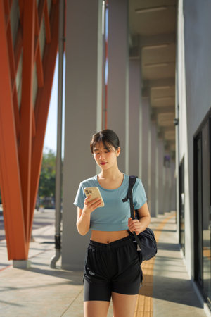 Young Asian woman walking after workout while checking smartphone outside gym building.