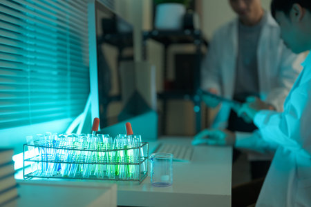 Test tubes with green solutions glow under laboratory light while scientists work in the background.