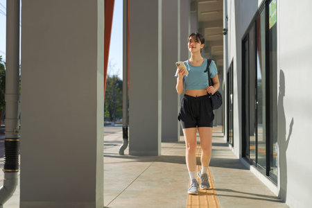 Young Asian woman walking after workout while checking smartphone outside gym building.の写真素材