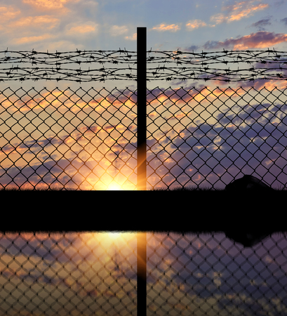 Concept of security. Silhouette of a metal fence with barbed wire on the background of sunsetの写真素材