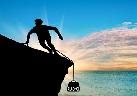 Concept of the disease of alcoholism. Silhouette of a man on a rock and alcohol in the form of cargo on sea sunset backgroundの写真素材