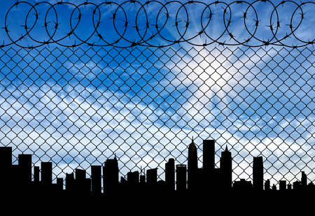 Concept of a border zone. Silhouette of the city behind a fence topped with barbed wire against a beautiful skyの写真素材