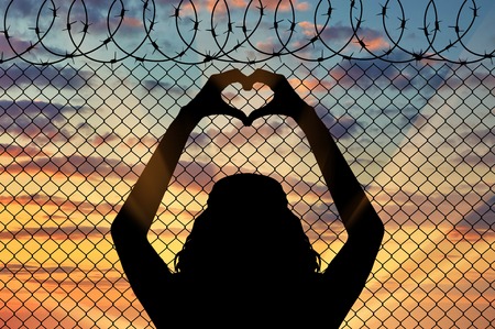 Concept of the refugees. Silhouette of a refugee shows a gesture from the hands of the heart on the background of the fence near the borderの写真素材