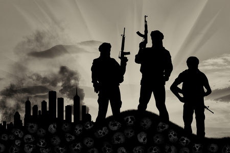 Concept of a terrorist attack. Silhouette of terrorists with a rifle standing on a pile of skulls on the background of the city in smokeの写真素材