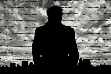 Refugees concept. Silhouette of refugee men near the border fence in the background of the urban landscapeの写真素材