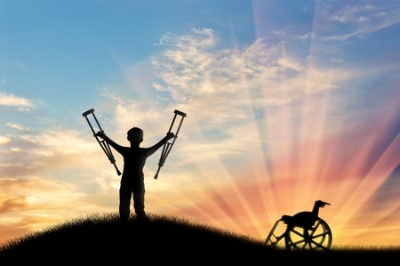 Happy child with crutches disabled person next to wheelchair on hill and sunset. Concept of disabilityの写真素材