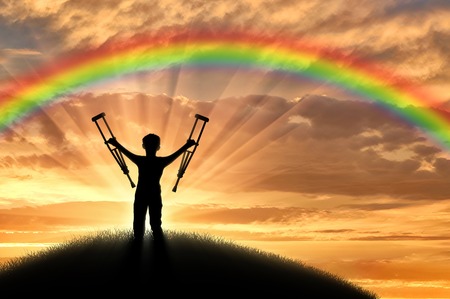 Child disabled with crutches on a hill on a background of a sunset rainbow. Concept of disabilityの写真素材