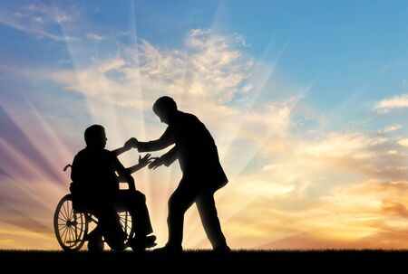 Man helps man in wheelchair to stand up on background sunset. Concept help disabled personsの写真素材