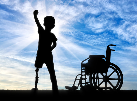Children with disabilities concept. Happy disabled boy with a prosthetic leg standing near a wheelchair against the skyの写真素材