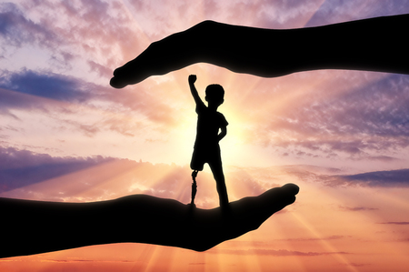 Help children with disabilities concept. Happy disabled boy with a prosthetic leg standing in the helping hands at sunsetの写真素材