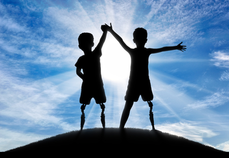 Children with disabilities. Two boys of a disabled person with a prosthetic leg standing, holding hands, on top of a hillの写真素材