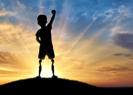 Children with disabilities concept. Happy disabled boy with a prosthetic leg standing on top of a hill at sunsetの写真素材