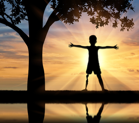 Children with disabilities concept. Happy disabled boy with a prosthetic leg standing near a tree at sunset and the river with its reflectionの写真素材
