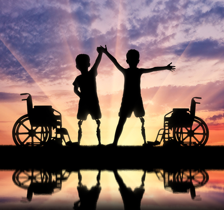 Children's disability concept. Two happy boy with a disability with a prosthetic leg standing near a wheelchair at the river with their reflectionの写真素材