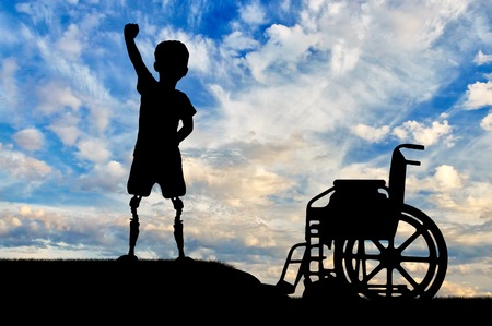 Children with disabilities concept. Happy disabled boy with a prosthetic legs standing near a wheelchair against the skyの写真素材
