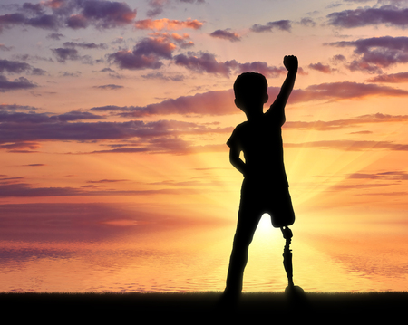 Children with disabilities concept. Happy disabled boy with a prosthetic leg standing at the sea at sunsetの写真素材