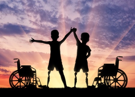 Children's disability concept. Two happy boy with a disability with a prosthetic leg standing near a wheelchair on sunset backgroundの写真素材