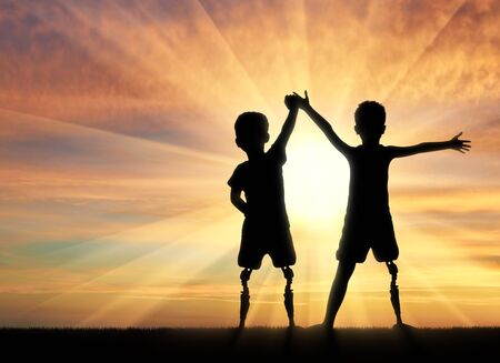 Children with disabilities. Two boys of a disabled person with a prosthetic leg standing, holding hands on sunset backgroundの写真素材