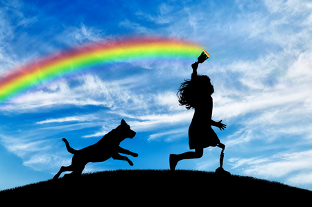 Children's disability concept. Child has a disability with a prosthetic leg with a brush in hand runs and draws a rainbow in sky with his dogの写真素材