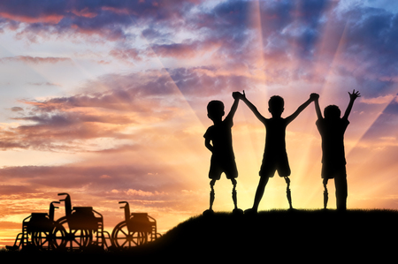 Children's disability concept. Happy children with disabilities with a prosthetic leg standing, holding hands at sunset, and wheelchairsの写真素材