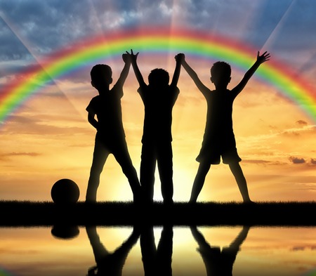 Childhood concept. Three happy children holding hands on a rainbow background and the sunset over the water with their reflectionの写真素材