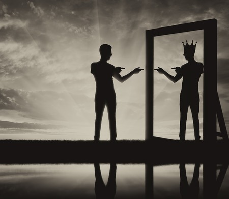 Concept of a narcissistic and egoistic man. Silhouette of a man standing, motivating himself at the mirror and sees in the reflection of himself with a crown on his headの写真素材