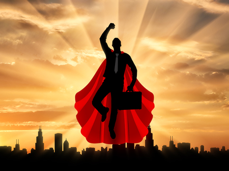 Superman businessman superhero. Silhouette of a superman businessman with a briefcase flying in the sky over the city at sunsetの写真素材