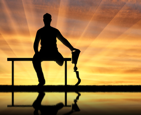 Silhouette of a man holding the hand of his prosthetic leg, sitting on a bench by the river with his reflection . The concept of people with prosthetic legsの写真素材