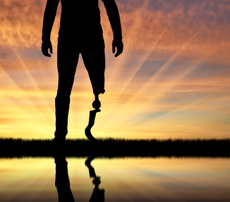 A man with a prosthetic leg standing at sunset close-up and its reflection in the water. The concept of a disabled person with a prosthetic legの写真素材