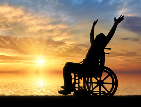 Happy disabled person in a wheelchair with arms raised on a background of sea sunset. The concept of happy people with disabilitiesの写真素材