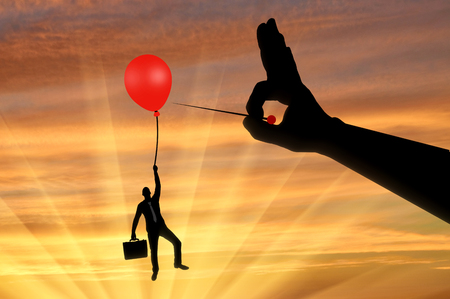 Silhouette of a man in the air clinging to a balloon and a hand with a needle wants to burst it. Concept of risks and complexities in businessの写真素材