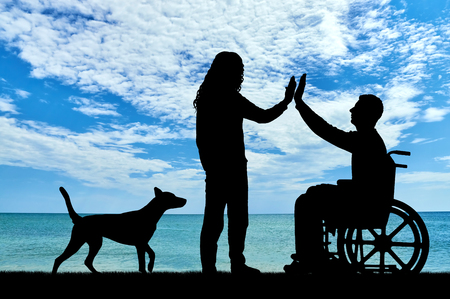 A man disabled in a wheelchair with having fun with friend and dog by the sea. The concept of support and assistance to people with disabilitiesの写真素材