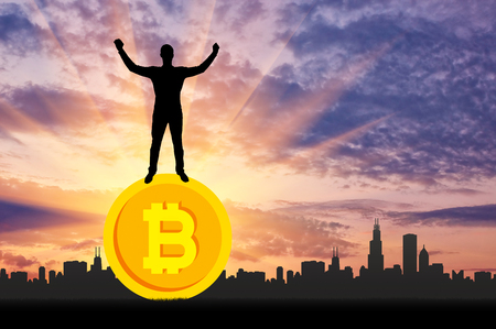 Happy man standing on a coin bitcoin against the background of the city. The concept of success in the crypto currencyの写真素材