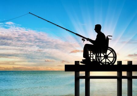 Silhouette of a disabled man in a wheelchair with a fishing rod in his hand fishing near the water on the pier. Concept of leisure and leisure of people with disabilitiesの写真素材