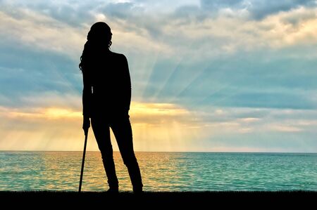 A silhouette of a woman supports herself with a crutch admiring the sea sunset. Concept of people in rehabilitationの写真素材