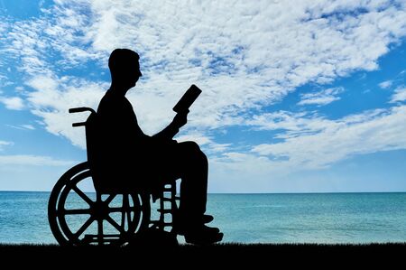 Silhouette of a disabled man in a wheelchair reading a book by the sea. Concept of the way of life of people with disabilitiesの写真素材