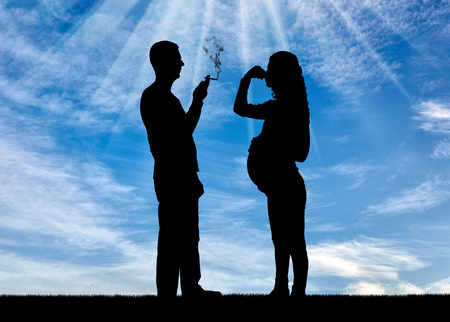 Silhouette of a man egoist smoking near a pregnant woman as she covers her nose with her hand from the smoke.の写真素材