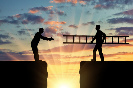 A silhouette of a man carries a ladder to another man who is on the other side of the abyss. The concept of teamwork and mutual assistanceの写真素材