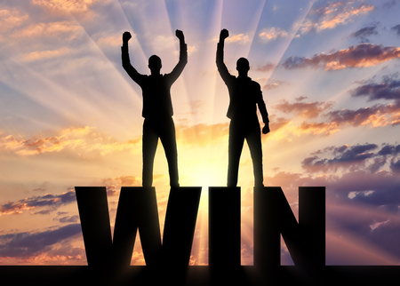 Silhouette of two happy men with raised arms standing on the word win. The concept of winning and mutual benefit of business partnersの写真素材