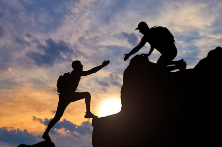 Silhouette of a male mountaineer working in a team with a partner, giving him a helping hand. The concept of a helping handの写真素材