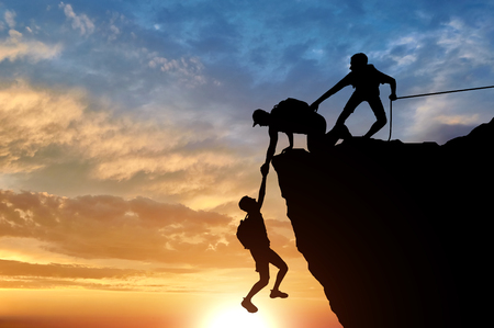 Silhouette of two male climbers rescuing another male climber pulling his arm. Conceptual scene climbers helpの写真素材