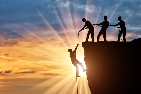 Silhouette of three climbers pulling out of the cliff of a man. Conceptual scene of male...