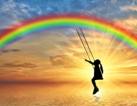 Silhouette, little girl child on a swing rainbow over the sea. Conceptual image of a happy childhoodの写真素材