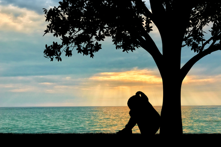 Silhouette of sad little girl sitting under a tree by the sea crying. Conceptual image of parents' divorceの写真素材