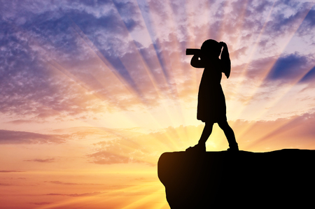 Silhouette of little girl child standing on top looking through binoculars. The concept of children's curiosity and discoveryの写真素材