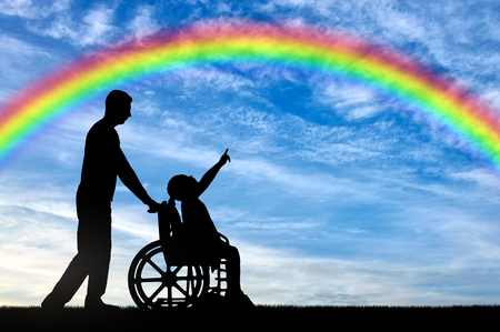 Silhouette of a disabled child girl sitting in a wheelchair and her dad showing a finger at a rainbow in the sky. Conceptual image of the life of children with disabilitiesの写真素材