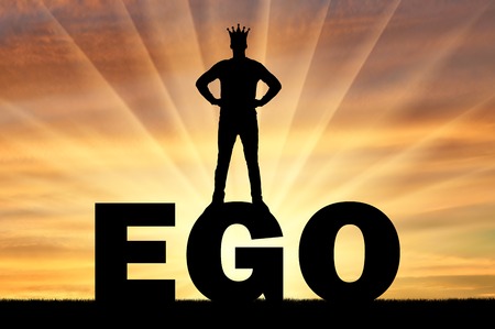 Silhouette of a man with a crown on his head standing on the word ego against the backdrop of a sunset. Concept of selfishnessの写真素材